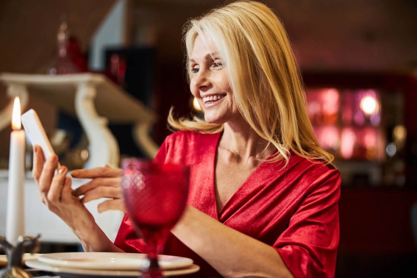 lächelnde, elegante blonde Frau im roten Seidenkleid schaut auf ihr Handy in einem Restaurant, um eine sugar mommy Schweiz finden zu können.