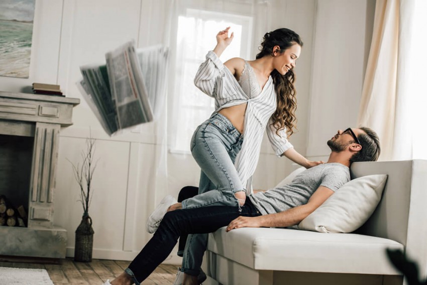 Leidenschaftliches Sexdate auf dem Sofa als Stressabbau, Frau in Dessous beugt sich &uuml;ber Mann, fliegende Zeitungen symbolisieren das Vergessen des Business-Alltags durch unverbindlichen Sex.