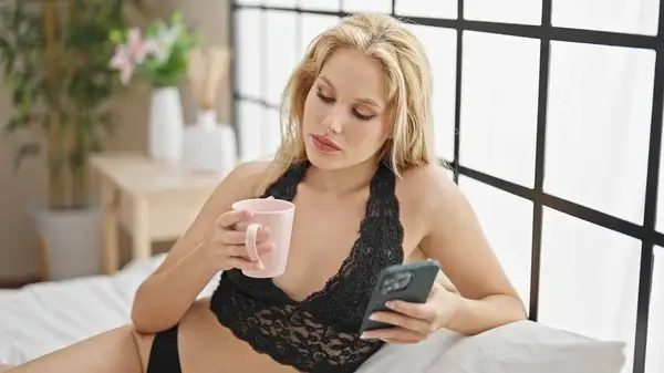 Sexting im Schlafzimmer – Blonde Frau in schwarzer Lingerie chattet sexy mit dem Handy bei einem heißen Sexdate.